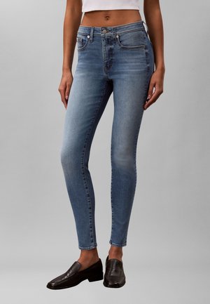 Femme portant un jean skinny bleu délavé, un top court blanc et des mocassins en cuir noirs, debout devant un fond gris uni.