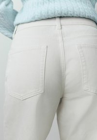 Osoba v krémových džínách z denimu a světle modrém texturovaném svetru, zobrazená zezadu od pasu po boky.