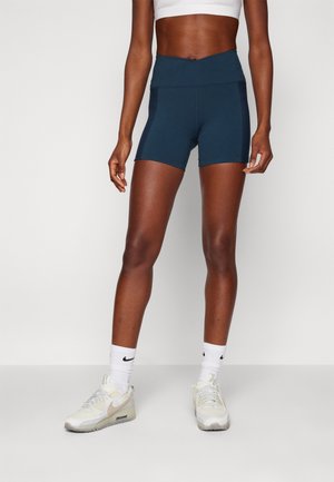 Personne athlétique portant un short cycliste bleu marine taille haute, un soutien-gorge de sport blanc, des chaussettes blanches Nike et des baskets Nike blanc cassé sur un fond uni.