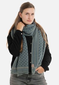 Lichtblauwe sjaal met een patroon en donkere accenten, voorzien van een geometrisch ontwerp; zachte textuur, gedragen over een zwarte geribbelde trui.