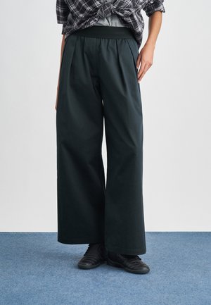 Person trägt lockere schwarze weit geschnittene Hosen, schwarze Sneakers, graues Shirt und gebundenes schwarz-weiß kariertes Hemd, steht auf blauem Teppich.