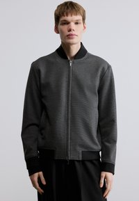 Μαύρο και γκρι μπομπέρ μπουφάν με φερμουάρ μπροστά, ριμπ μανσέτες και στρογγυλή λαιμόκοψη. Το υλικό έχει υφή με ανάγλυφη επιφάνεια.