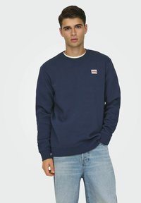Mørkeblå sweatshirt lavet af en blanding af bomuld, med rund halsudskæring, ribbede manchetter og et lille rektangulært logo-patch på brystet.