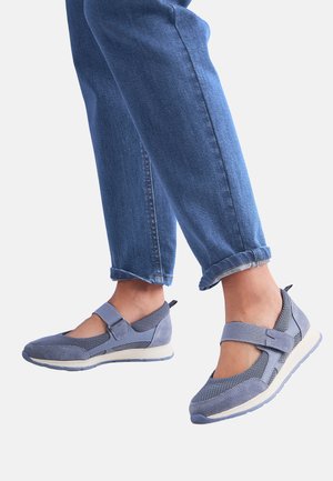 Person trägt blaue Mesh- und Wildleder-Mary-Jane-Sneaker kombiniert mit hochgekrempelten blauen Jeans vor weißem Hintergrund.