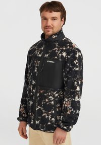 Fleece-Jacke mit schwarz-weißer abstrakter Musterung, kontrastierender schwarzer Reißverschlusstasche auf der Brust und hohem Kragen für Wärme und Stil.