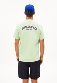 Lichtgroene t-shirt met "ARMEDANGELS" en "SINCE 2007" in marineblauw gedrukt op de achterkant; gedragen met donkere shorts en een blauwe pet.