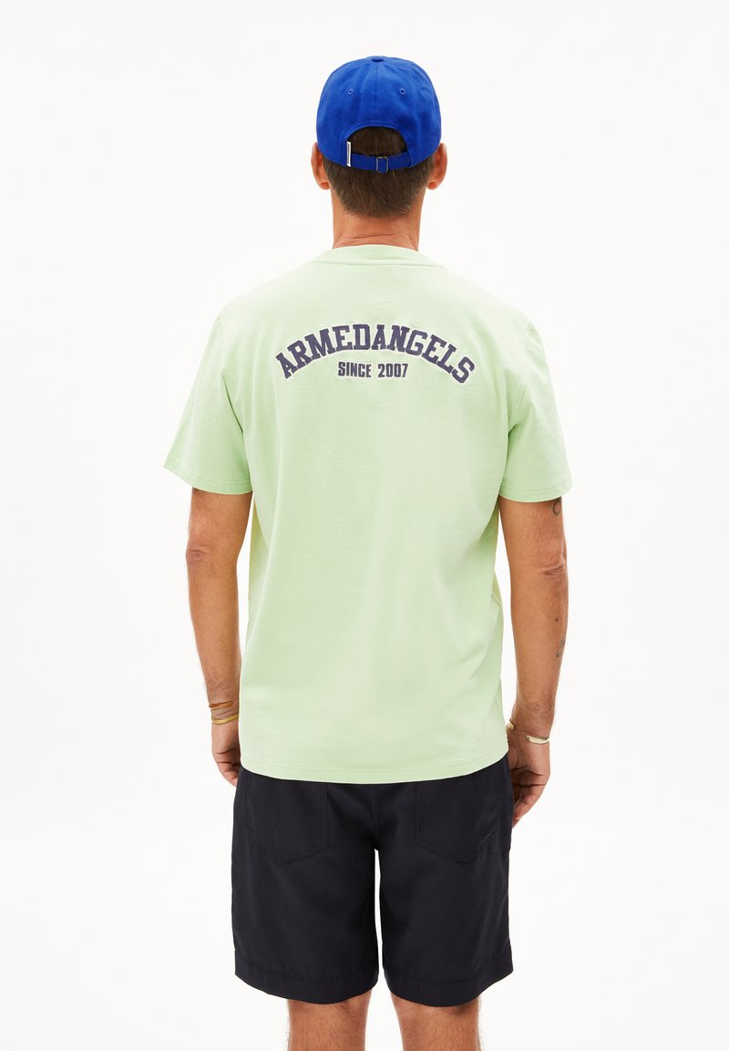 Lichtgroene t-shirt met "ARMEDANGELS" en "SINCE 2007" in marineblauw gedrukt op de achterkant; gedragen met donkere shorts en een blauwe pet.