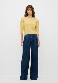 Maglione giallo lavorato a maglia con un motivo testurizzato e decorazioni floreali sul colletto, abbinato a jeans in denim scuro a vita alta e gamba ampia.