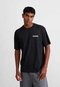 T-shirt nera in cotone con collo a giro, maniche corte e stampa bianca del logo sul lato sinistro del petto, abbinata a pantaloni corti grigi.