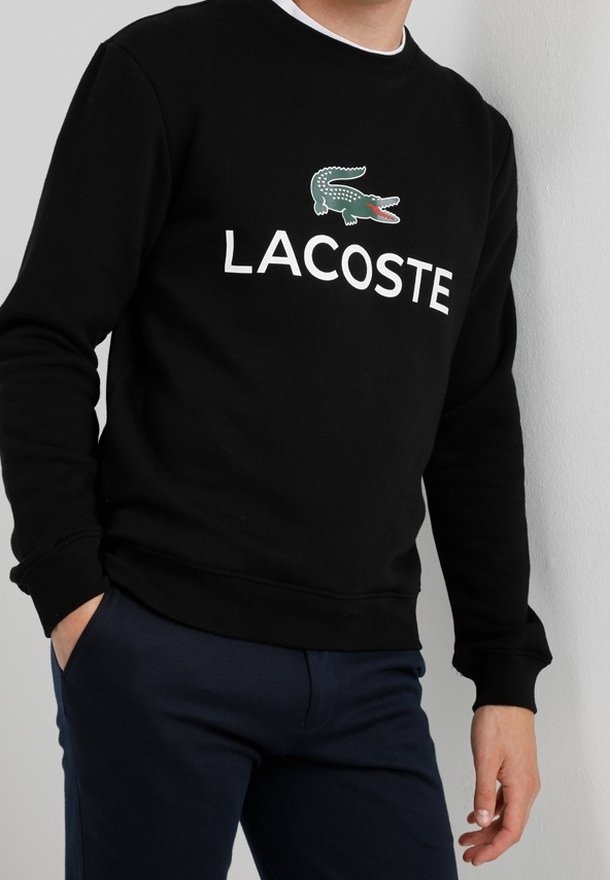 Černá mikina vyrobená z bavlny, která má zelené a červené logo krokodýla a bílé nápis "LACOSTE" přes hruď. Klasický kulatý výstřih.