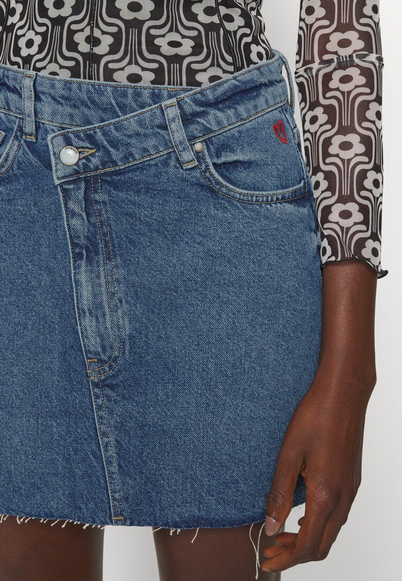 Gonna mini in denim con orlo sfrangiato, lavaggio blu medio, tasche frontali e ricamo a forma di cuore rosso sulla vita, abbinata a una maglia a fantasia.