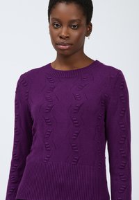 Junge schwarze Frau, die einen strukturierten lila Strickpullover mit geripptem Saum und rundem Halsausschnitt trägt und vor einem schlichten, hellen Hintergrund steht.