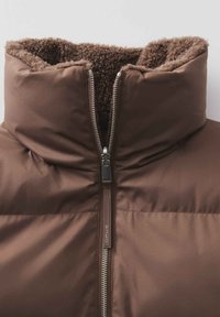 Braune Daunenjacke mit einem flauschigen braunen Fleecekragen, voller Vorderreißverschluss und glattem Außenmaterial, das horizontale Steppdetails aufweist.