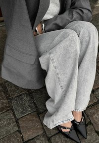 Jeans anchos grises con una textura deslavada, combinados con zapatos negros de punta con detalles de tiras. Un blazer gris está apoyado sobre las piernas.