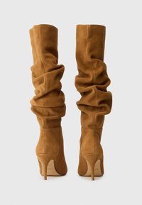 Bottes en suédine marron au-dessus du genou avec un design relâché, bout pointu et talon aiguille, présentant une texture lisse et des coutures discrètes.