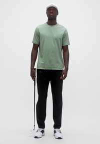 Άνδρας με ανοιχτό πράσινο μπλουζάκι, μαύρο παντελόνι, λευκό καπέλο και αθλητικά παπούτσια, στέκεται κρατώντας ένα μπαστούνι γκολφ σε λευκό φόντο.