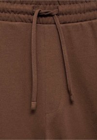 Pantalons de jogging en tissu brun avec une taille élastique et un cordon de serrage à l'avant, mettant en avant des coutures détaillées et une texture douce.