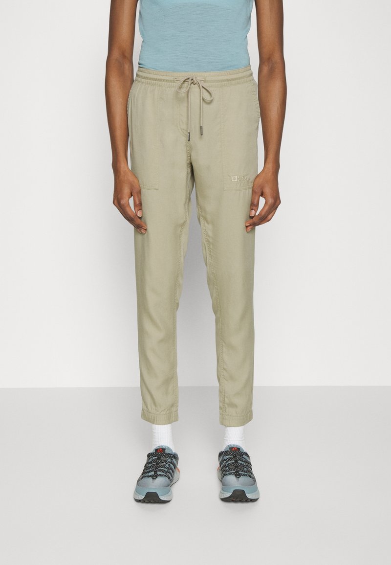 Ljusbeige cargobyxor med dragsko i midjan, sidofickor och elastiska muddar, kombinerat med blå träningsskor och vita strumpor.