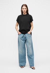 Kvinde stående med hænderne i lommerne iført sort kortærmet top, løstsiddende lyseblå jeans og sorte åbne tå-hæle på hvid baggrund.