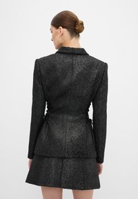 Blazer nero texture con design aderente, caratterizzato da una vita accentuata e un fondo svasato. Include un sottile dettaglio posteriore con lacci.