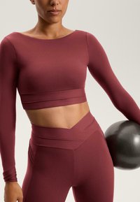 Burgundy hosszú ujjú crop top, amelyet magas derékú leggingsszel párosítottak, amelynek keresztbe fonott kialakítása van. Az anyag simának és rugalmasnak tűnik.