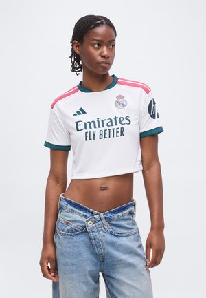 Młoda kobieta z zaplecionymi włosami, ubrana w krótki futbolowy T-shirt Realu Madryt i rozpięte niebieskie dżinsy, patrząca na bok.