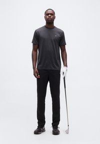 Άνδρας με μαύρη ενδυμασία γκολφ, κρατώντας μπαστούνι γκολφ με λευκό γάντι στο δεξί χέρι, στέκεται μπροστά σε απλό λευκό φόντο.