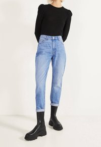 Haut noir côtelé à manches longues, jean bleu clair taille haute avec revers, et bottines noires épaisses. La tenue combine différentes textures.