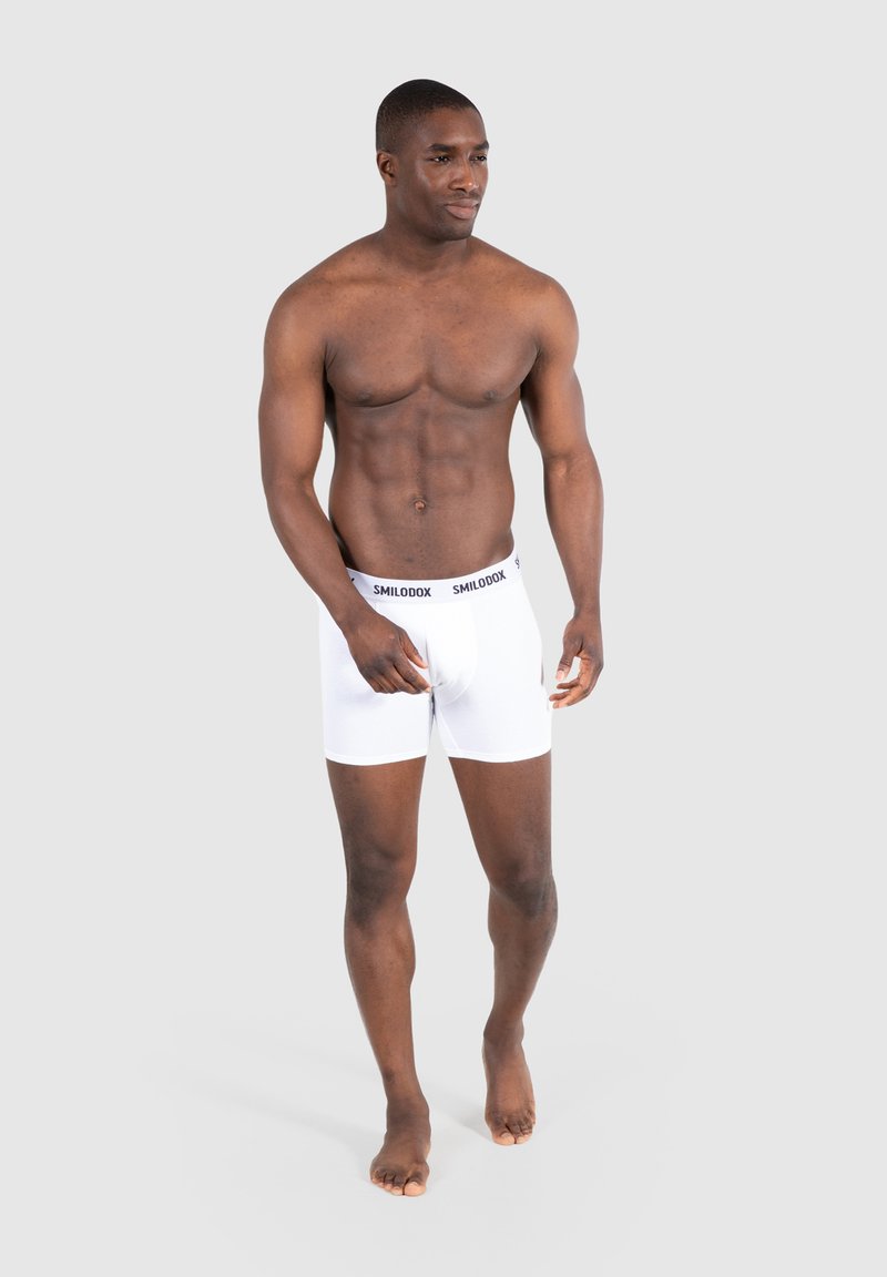 Boxer shorts bianchi in cotone con elastico nero a marchio, design aderente e texture liscia, indossati da una persona con una corporatura muscolosa.
