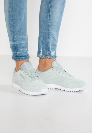 Jasnoniebieskie zamszowe sneakersy z białą podeszwą noszone z wyblakłymi, postrzępionymi, obcisłymi niebieskimi jeansami na białym tle.