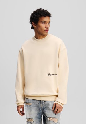 Bež dukserica s okruglim izrezom izrađena od mekog materijala, s rebrastim manžetama i donjim rubom, te logotipom "KARL LAGERFELD JEANS" u crnoj boji na dnu.