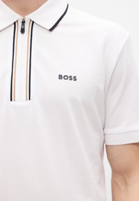 Λευκό πόλο μπλουζάκι με κλείσιμο με φερμουάρ στο μισό, μαύρες και χρυσές ρίγες, και μαύρο λογότυπο "BOSS" στο στήθος. Μαλακό, υφασμάτινο με υφή.