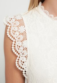 Balta mežģīņu topa ar sarežģītām ziedu rakstu detaļām, viļņainām malām un smalku izšuvumu gar plecu. Kopumā gluda tekstūra.