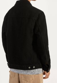 Svart denimjacka med krage, sidofickor och knapppynt vid ärmarna. Buren över en mönstrad ljus topp och beige byxor.