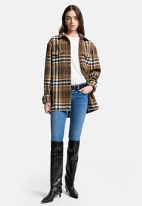 Bruin geruite flanellen shirt met zwart en witte accenten, knoopsluiting aan de voorkant, gecombineerd met lichtblauwe skinny jeans en zwarte kniehoge laarzen.