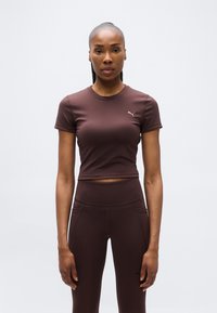 Γυναίκα που στέκεται με το πρόσωπο προς τα εμπρός, φορώντας εφαρμοστό κοντομάνικο crop top και ψηλόμεσα κολάν αθλητικού ρουχισμού σε σκούρο καφέ χρώμα.