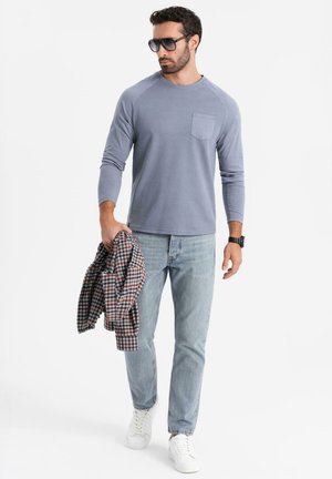 Helles blaues Langarmhemd mit einer Brusttasche, kombiniert mit hellen Jeans und weißen Sneakers. Hält ein kariertes Hemd. Schwarze Sonnenbrille.