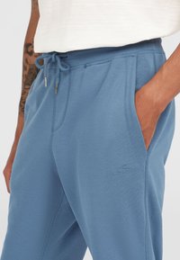 Blue sweatpants made of soft fabric, featuring an elastic waistband with drawstring, side pockets, and a logo detail on the left thigh.