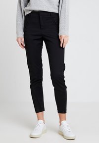 Svarta, slim-fit byxor med en slät textur, med sidofickor och en avsmalnande ben. Kombinerat med vita sneakers.