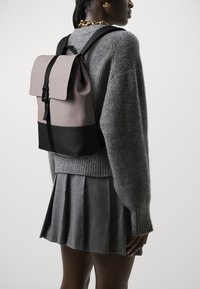 Backpack featuring a mauve upper section with a flap and black bottom. Made from smooth material, with adjustable straps and a simple design.