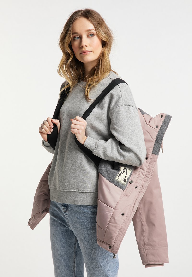 Eine hellrosa Jacke mit grauen Akzenten, quilted Textur und einem Logo-Patch, gehalten über einem grauen Sweatshirt, kombiniert mit blauen Jeans.