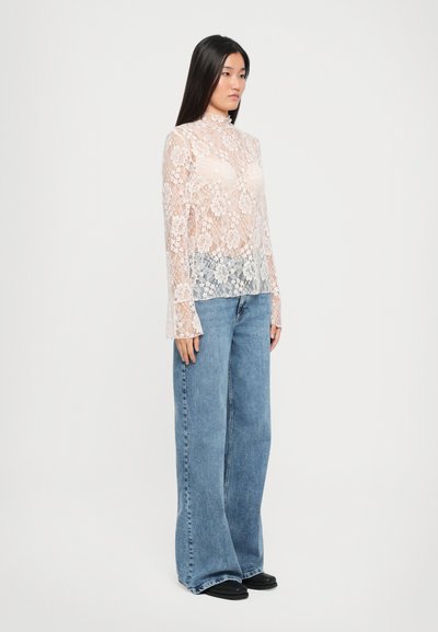 Lange mouw top van kant in lichtroze met bloemenpatroon en hoge hals, gecombineerd met losse blauwe denim jeans. Zwarte platte schoenen.