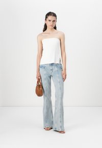 Hvit stroppeløs topp, lyseblå vide jeans med frontlommer, og en brun håndveske; modellen har på seg brune sandaler; minimal bakgrunn.