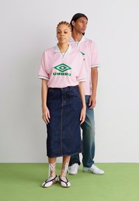 Duas modelos vestem conjuntos coordenados: uma camisa rosa da UMOBRO com detalhes brancos, uma saia de ganga e sapatos com atacadores em preto e branco.