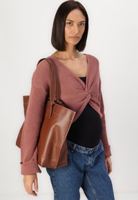 Braune Leder-Tote mit glänzender Oberfläche und kontrastierenden Nähten. Kombiniert mit einem geknoteten, rosafarbigen Strickpullover und Jeans.