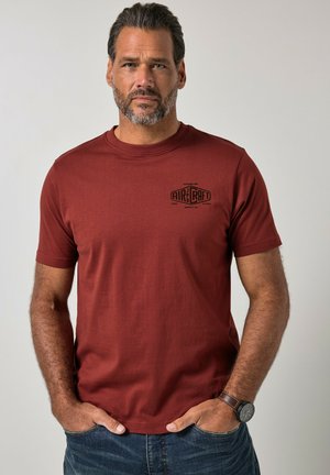 Mann mit kurzen grauen Haaren und Bart trägt ein rostfarbenes T-Shirt mit „Aircraft“-Logo, blaue Jeans und eine Armbanduhr, steht mit den Händen in den Taschen.