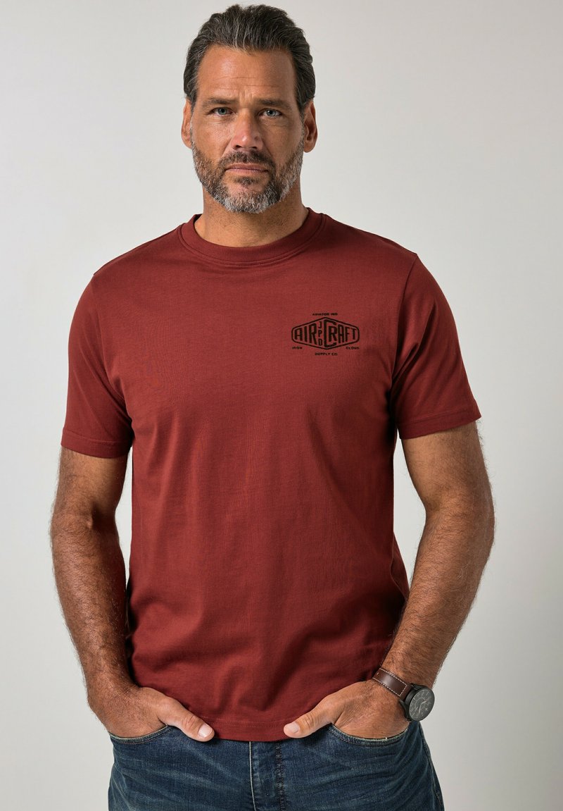 Homme aux cheveux gris courts et barbe porte un t-shirt couleur rouille avec le logo « Aircraft », un jean bleu et une montre-bracelet, debout les mains dans les poches.