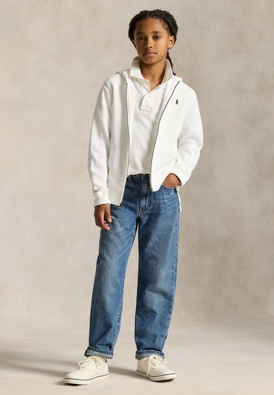 Enfant debout, main dans la poche, portant un sweat à capuche blanc zippé, un polo blanc, un jean bleu et des baskets blanches sur un fond uni.