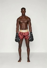 Pantalones cortos de boxeo de satén color burdeos con una cinturilla elástica dorada, que presenta un logotipo. Guantes de boxeo negros con acentos blancos. Postura descalza.