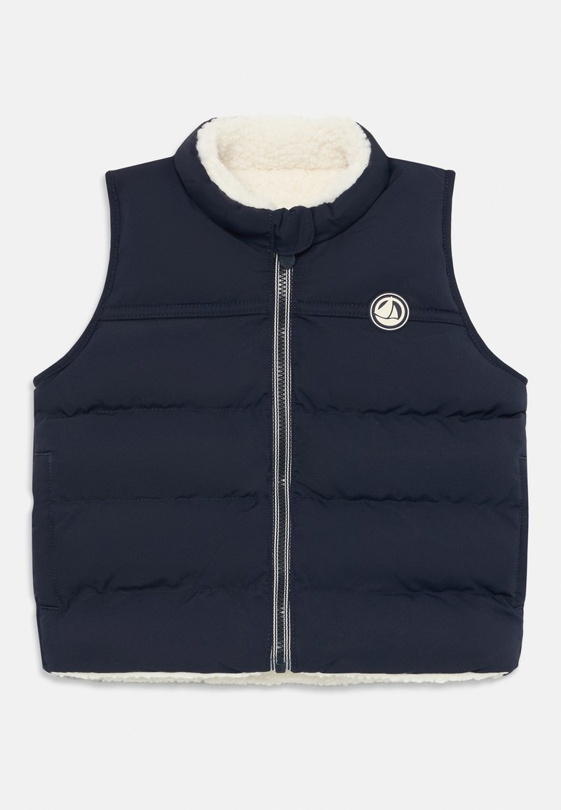 Marinblå vadderad väst med hög krage, försedd med en mjuk vit fleece-foder och en framdragkedja. Inkluderar en rund logotypdetalj på bröstet.